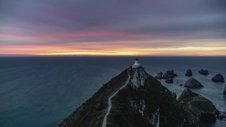Landscape of a rocky coast surrounded by the sea with a light house on the cliff against a reddish sky