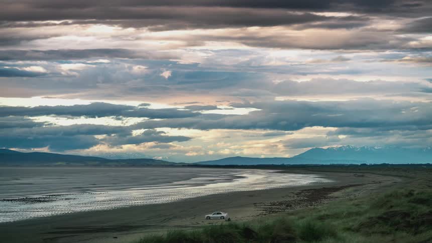 Beach landscape at dusk with a cloudy sky reflected in low tide water and a white car parked in the distance on the sand