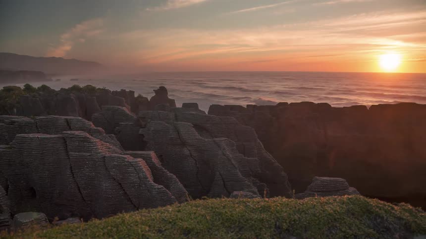 Coastal landscape at dusk with rock formations in the foreground and a foggy ocean in the background under a setting sun