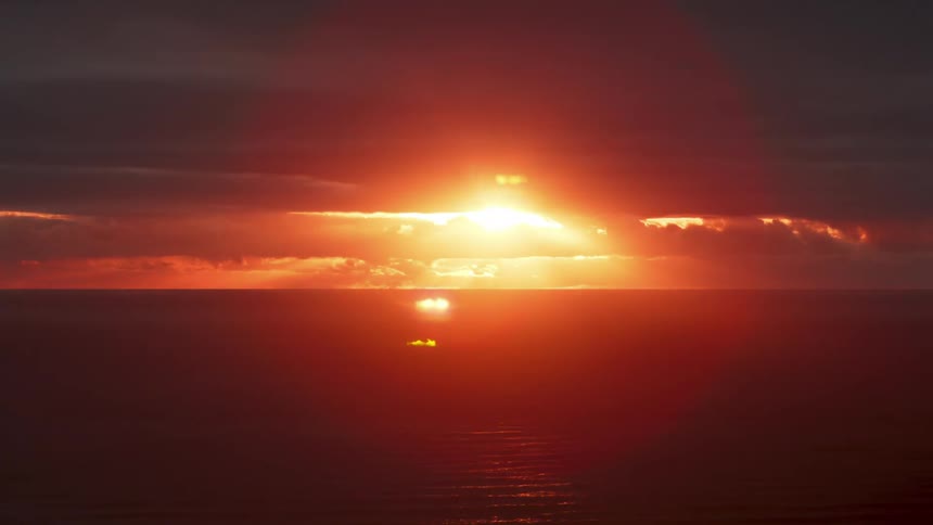 Setting sun flares orange then red among the cloudsover the ocean on the horizon