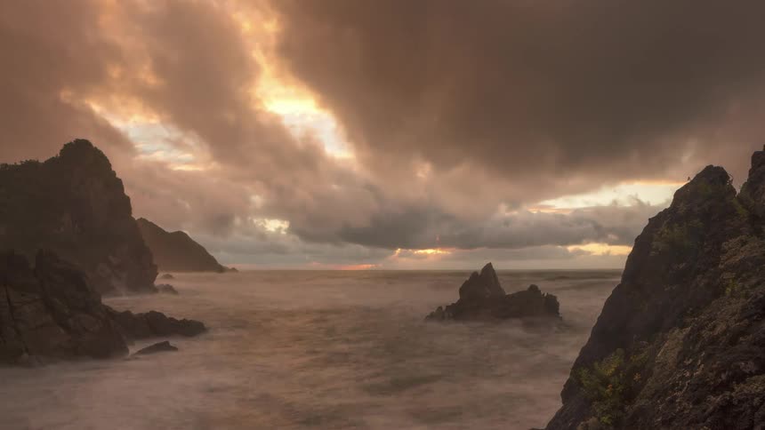 Dark clouds flow over waves crashing against a rocky shore