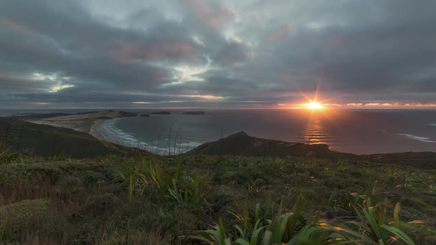 Heavy clouds flow over the shore as the setting sun flares red on the horizon
