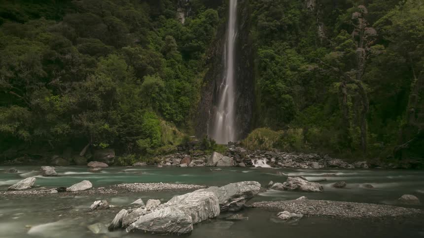 Time lapse of a slender waterall descending into a river under a cloudy blue sky