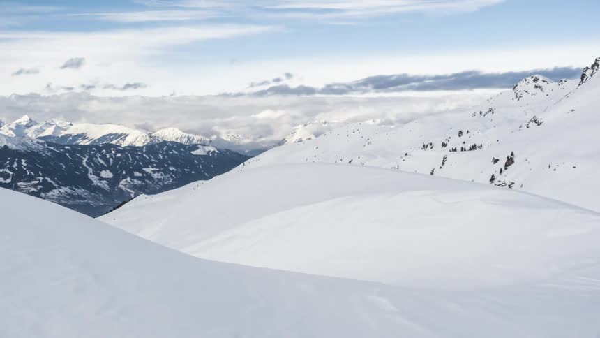 A mountain with lots of snow covering it and several white blouses in the sky moving to the right