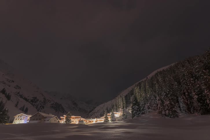 A small town of houses in a region with a lot of snow and trees and it is getting dark and the houses turn on the lights