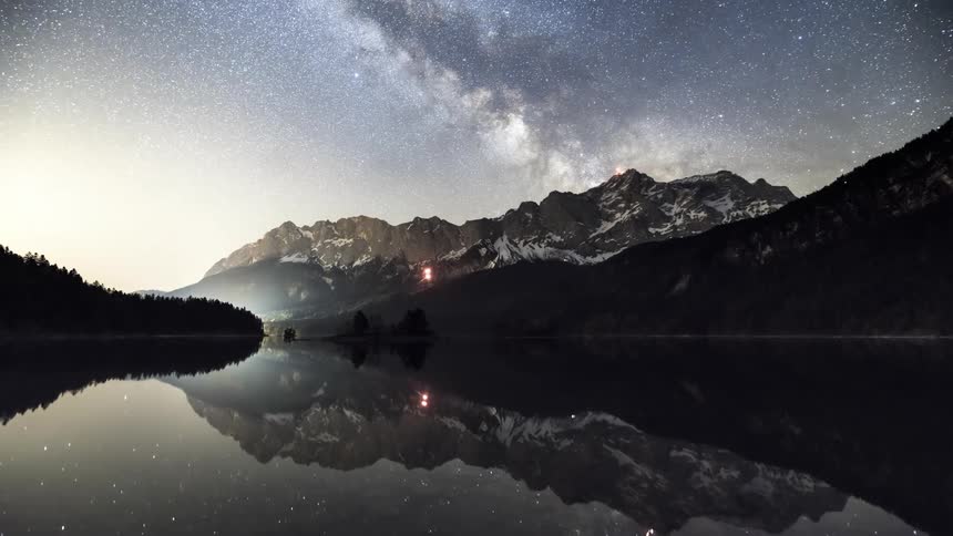 Calm lake reflecting snowy mountains and a forest under a dawning starry sky