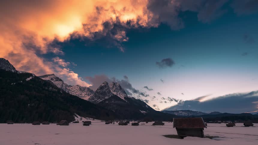 People walk along the paths through a snow covered town near the mountains as night falls