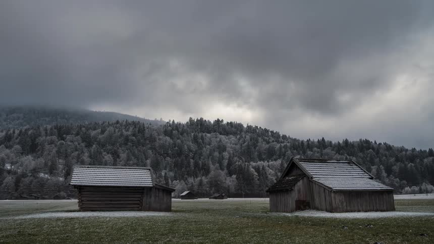 Two wooden houses in a field surrounded by mountains and conifers on a cloudy day
