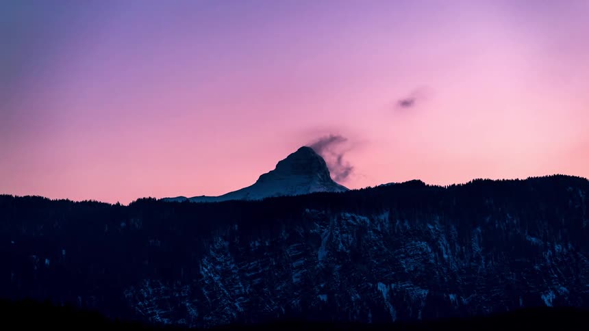 Tall mountain towering over forested mountainous terrain stands silhouetted against a pink sky