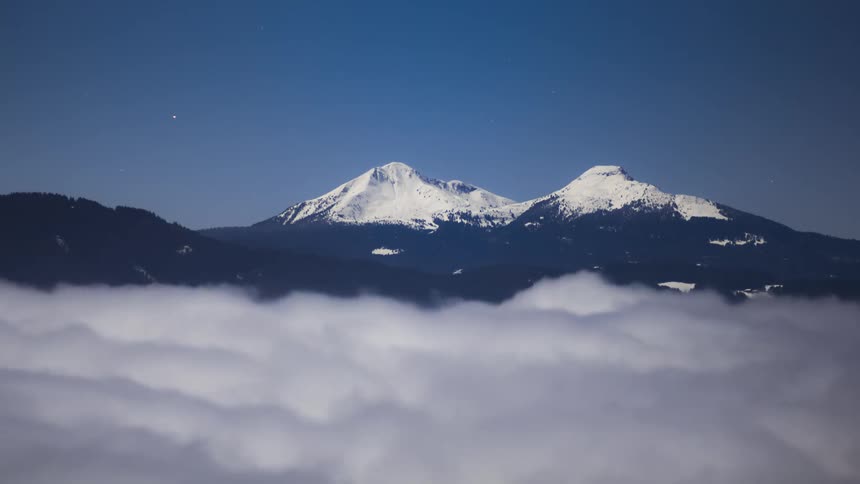 Several white clouds moving quickly and in the background the peak of two mountains covered in snow