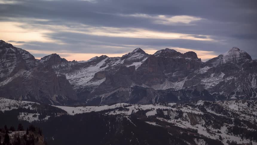 Clouds flow through the red tinted sky over snowy mountains with forested lower slopes