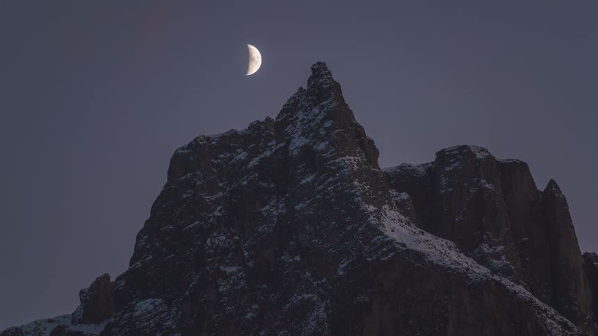 A half moon moves across the sky over a mountain peak
