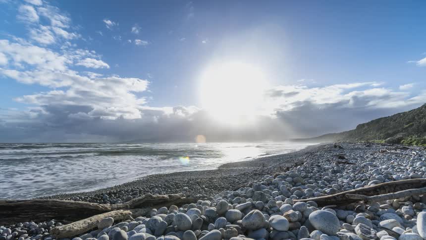 Pebble beach with gentle waves on the shore and a tree trunk in the sand under a partly cloudy sky with dense clouds looming