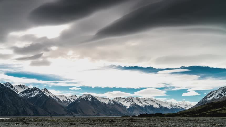Dense clouds cross the sky over a valley between snowy mountains