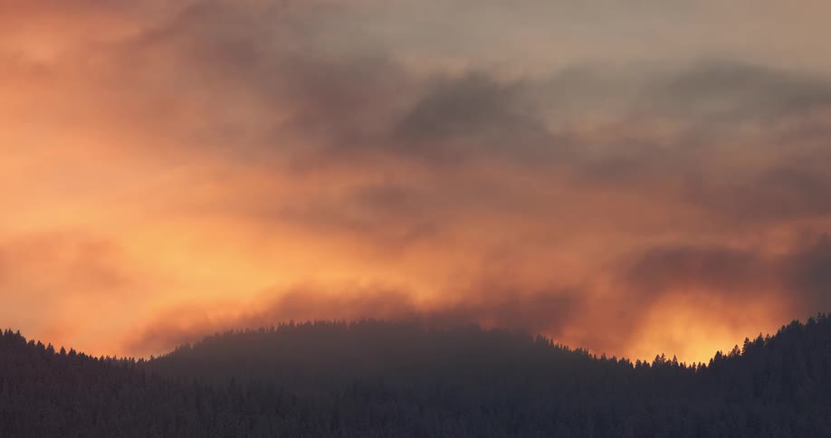 A warm orange and yellow sunset sky mist and clouds over a mountain with forests