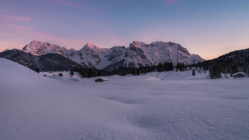 Landscape of mountains and conifers covered in snow with the sky turning purple
