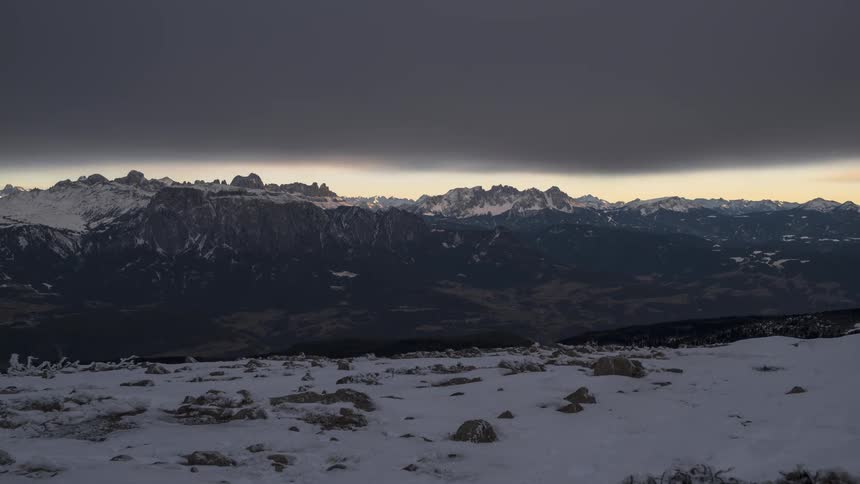 Snow covered plateau overlooks dark mountainous terrain as the sun sets under an overcast sky