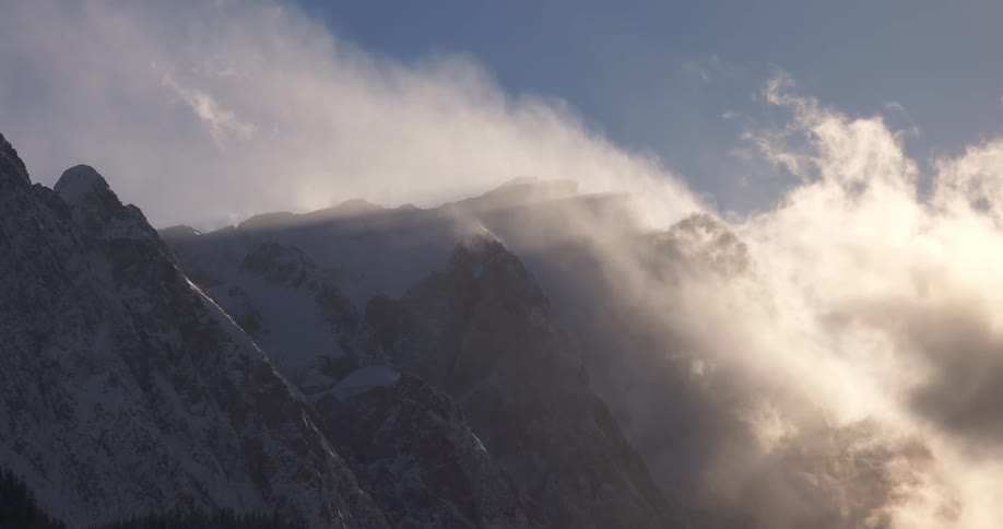 The peak of a mountain range covered in snow with a light breaking through white clouds passing over the mountains