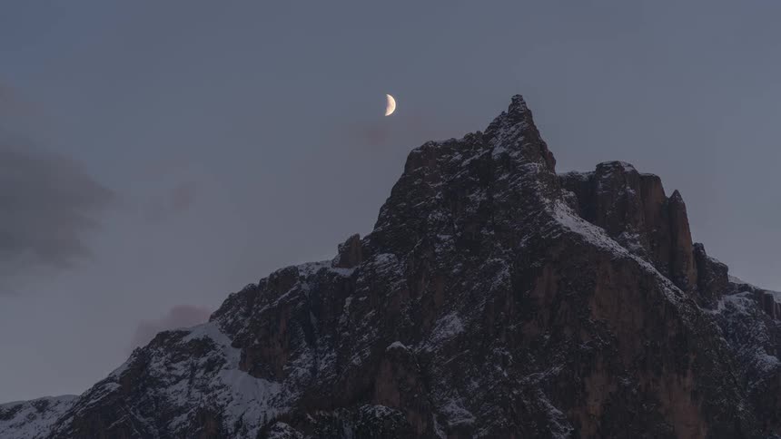 Crescent moon travels through the sky over a snow dusted mountain in winter during the day and then the night