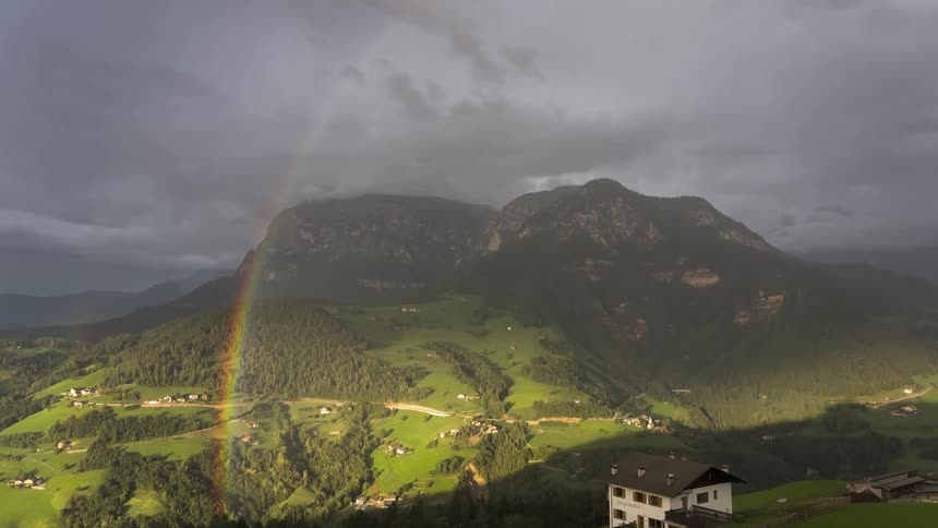 Mountainous landscape under a cloudy sky with sunlit areas and a rainbow