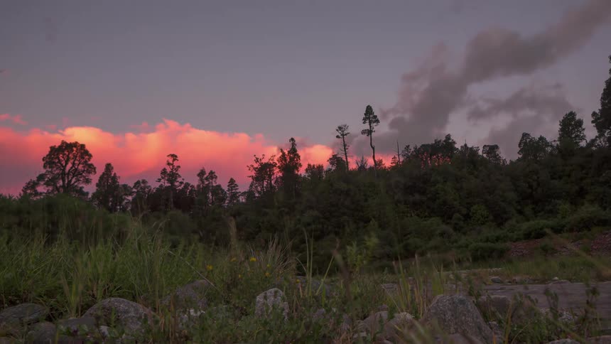 A region with rocks and vegetation and some trees behind and the sky with orange clouds is getting dark and turning to gray clouds