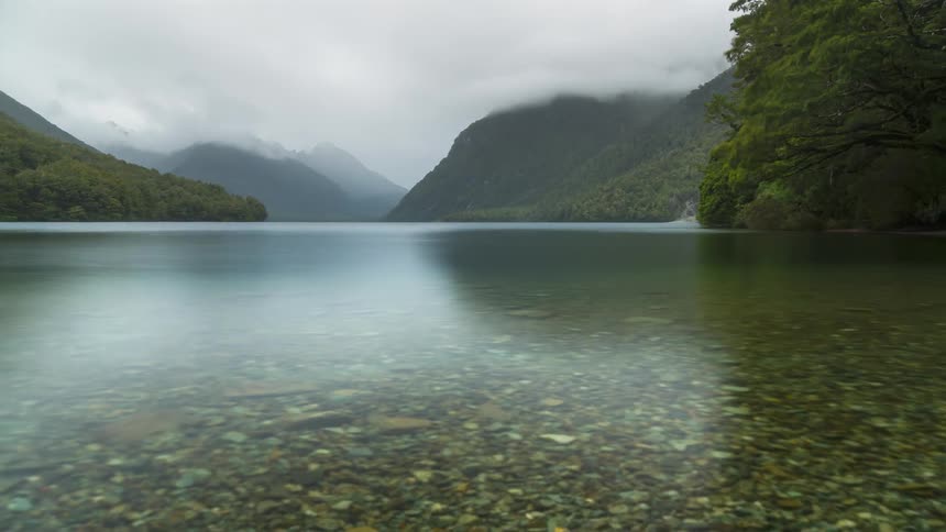 Tranquil lake with clear water surrounded by mountains and greenery under a cloudy sky