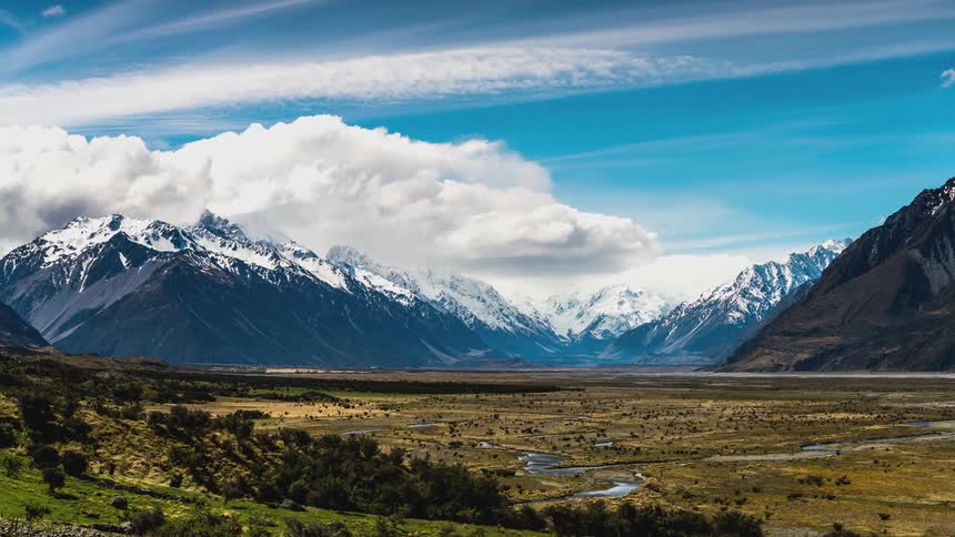 Clouds passing over a grassy plain and a mountain range with snow covered peaks