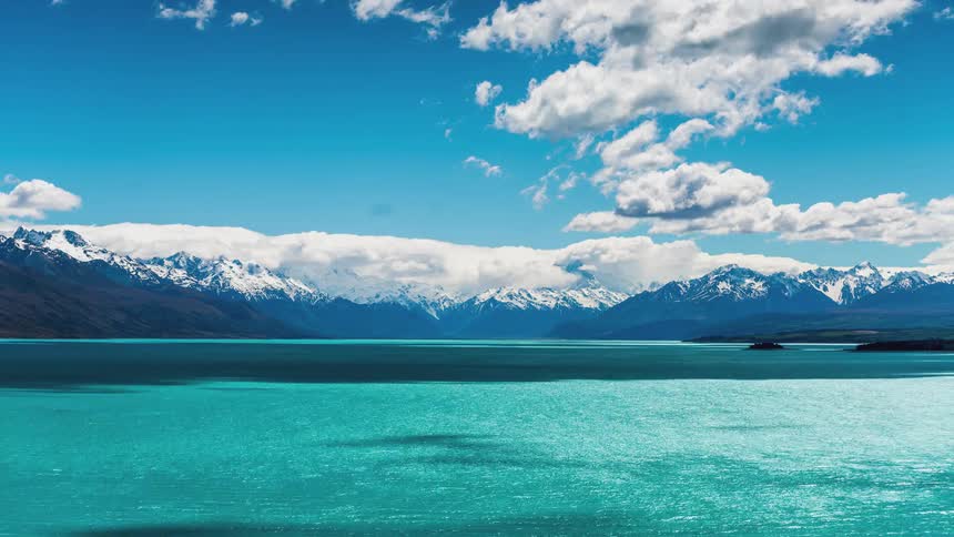 Crystal clear blue lake surrounded by snowy mountains in clouds passing by
