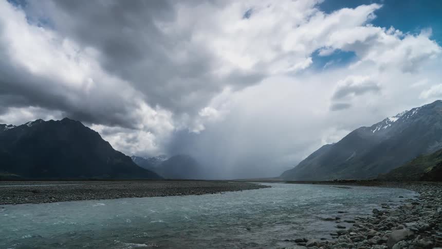 Pale blue waterway flows between rock covered banks passing foggy mountains on a cloudy day