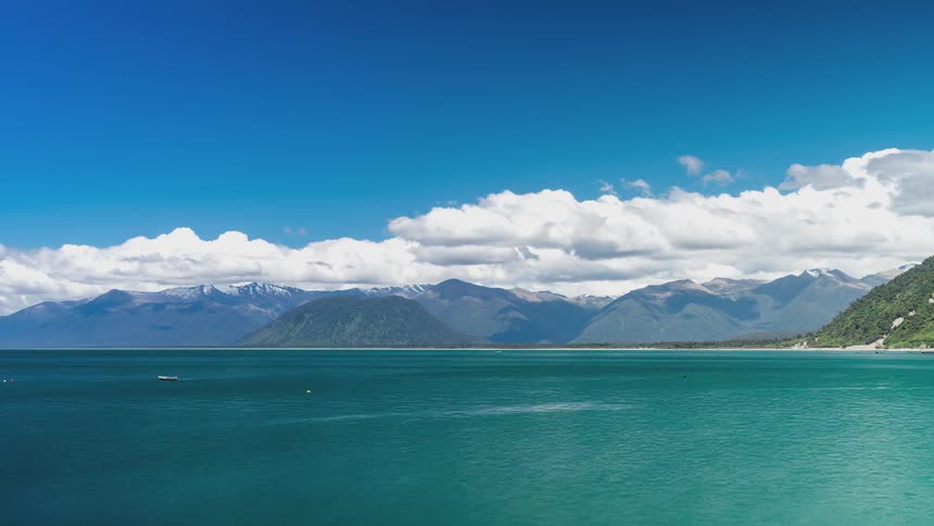 Vast turquoise lake with a strip of land and mountains in the background under a clear blue sky with clouds