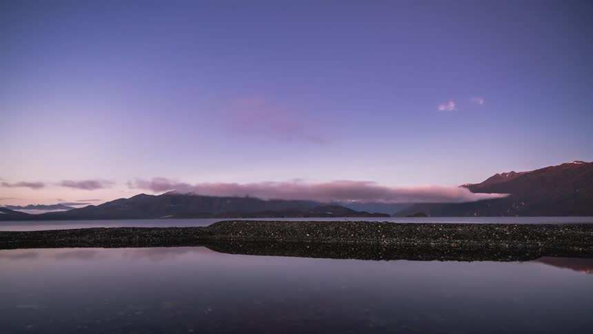 Calm lake at dawn with mountains in the background under a sky that changes color