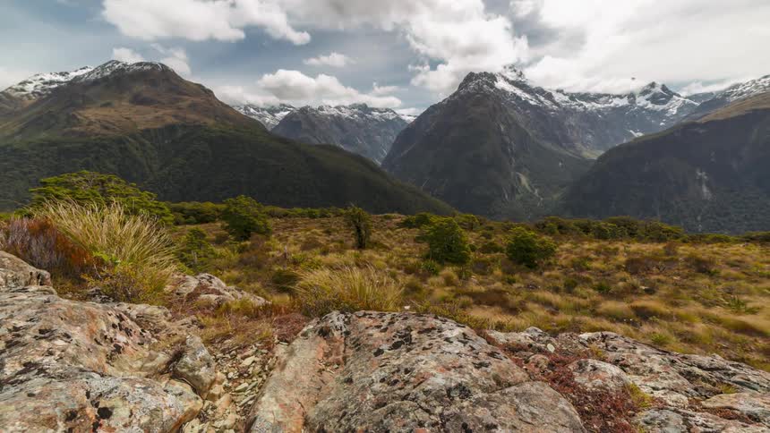 Orbiting time lapse of a rocky terrain next to a large mountain range under rolling clouds