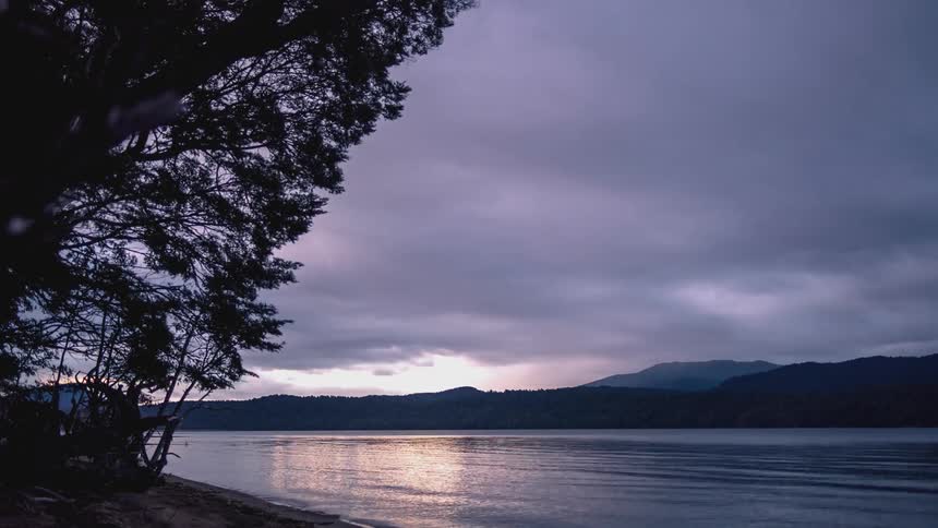 A region where there is a lake surrounded by mountains in the background and in the sky there are many white and gray clouds moving in the evening