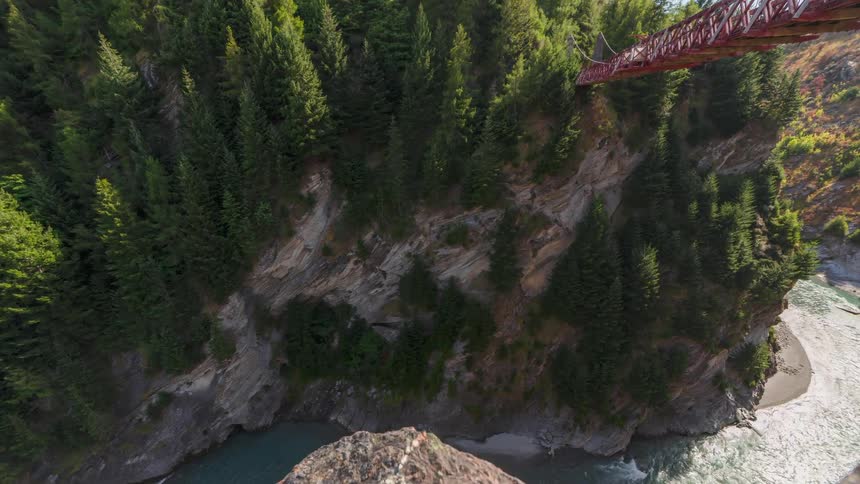 A red bridge stretches across a deep river gorge on a sunny day