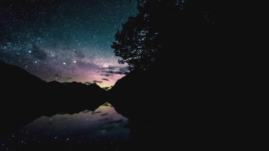 Starry night sky with shades of blue and the silhouette of trees and mountains on the shore of a lake