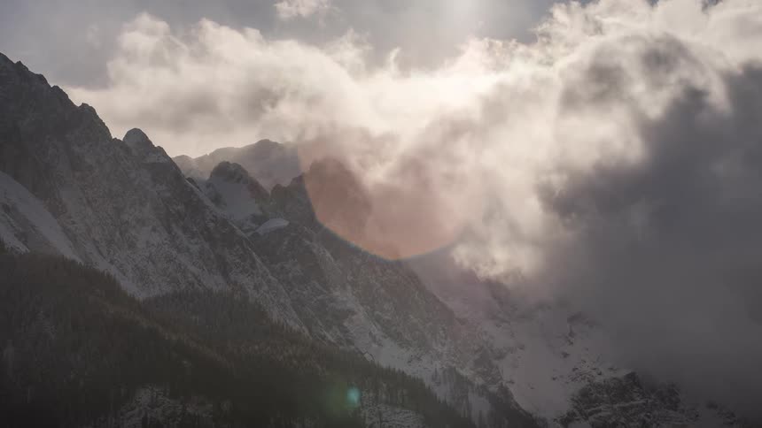 Light glares as clouds pass over a snowy ridge and forested slopes