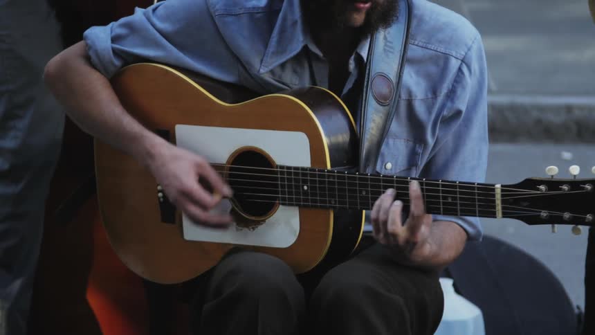 A man sitting outside playing guitar while people walk by