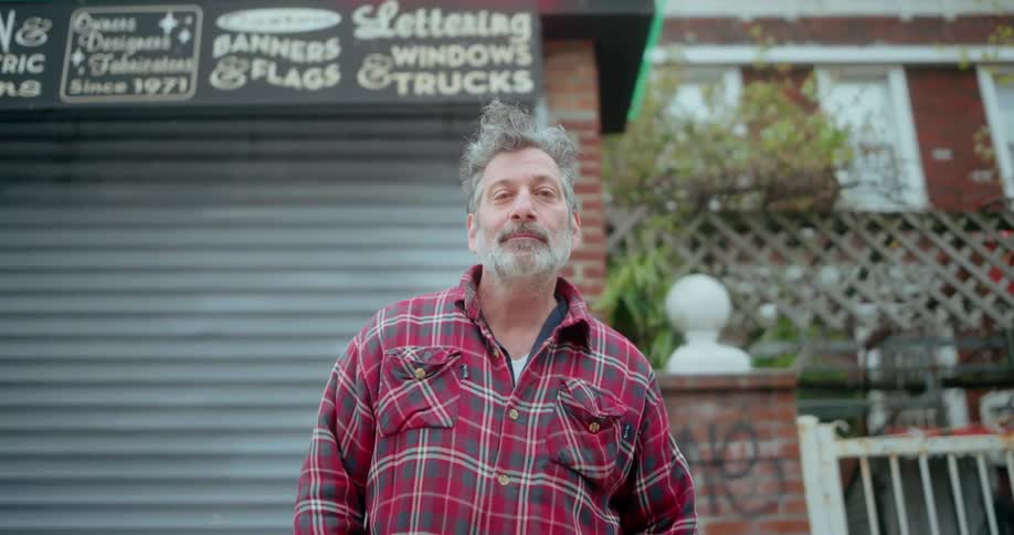 A male shop owner stands proudly in front of his neon shop