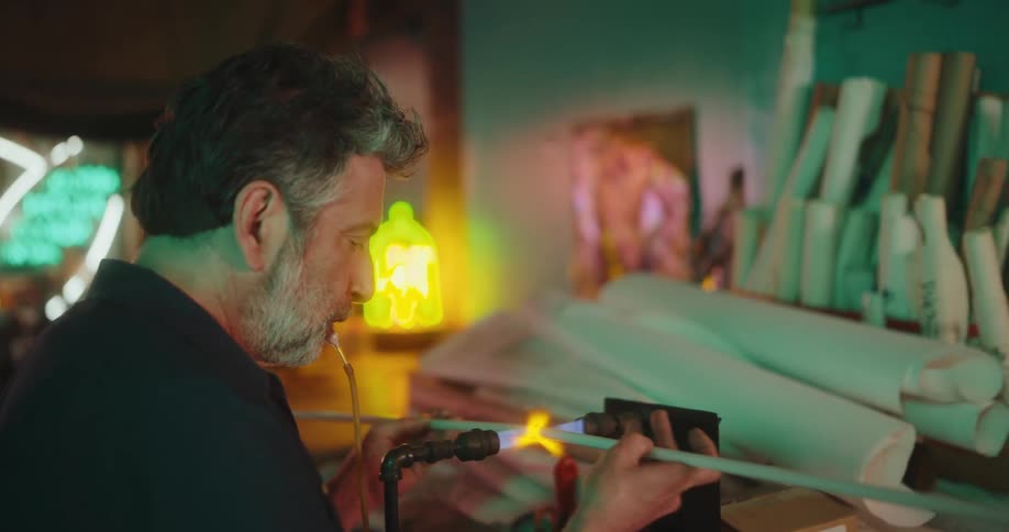 Man working with a blowtorch with a glass tube in a workshop surrounded by papers and boxes