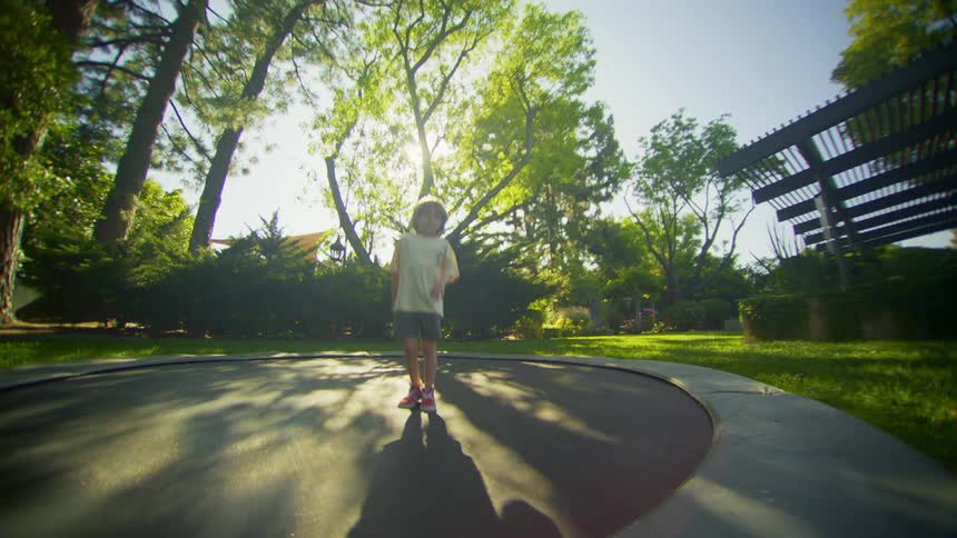 A young boy jumps on a trampoline as his sister runs past on the grass
