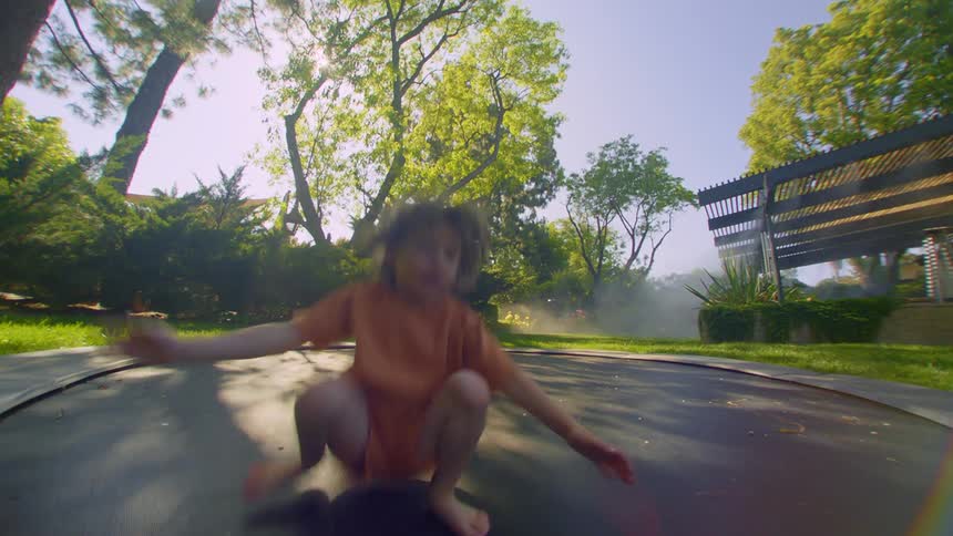 A little boy in orange clothes is jumping and it is a trampoline that is in a yard with several sunshine trees
