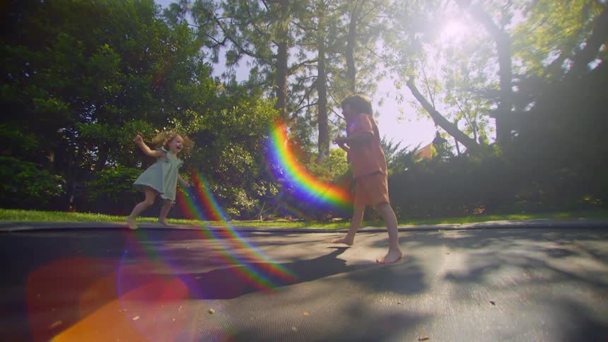 A little boy and a little girl jumping on a black trampoline and behind there are several trees and bright sunlight