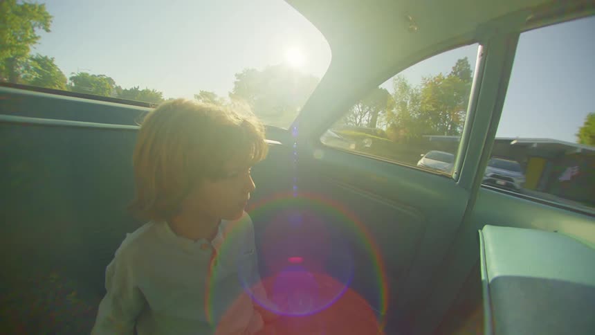 A young boy sits in the back seat of a car as it drives down a street
