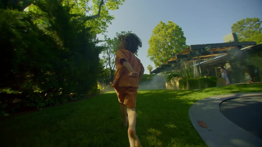 A little boy has his back turned and is running on a lawn in a backyard