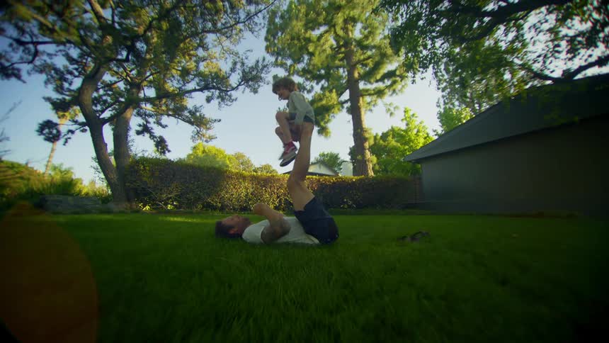 Father lying on the grass playing with his son who is sitting on his fathers feet in an outdoor setting