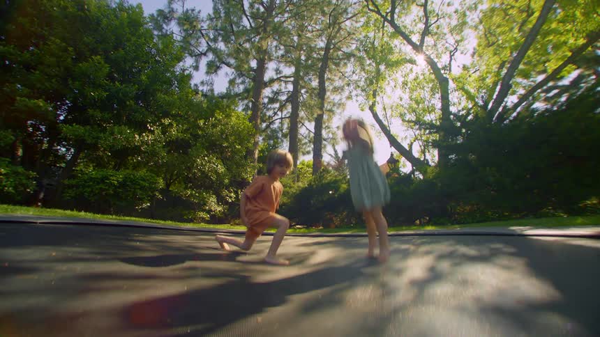 Two children playing jumping on the trampoline in a backyard surrounded by trees