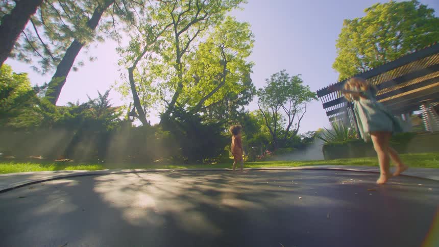 Two kids jump around on a large ground trampoline in a backyard