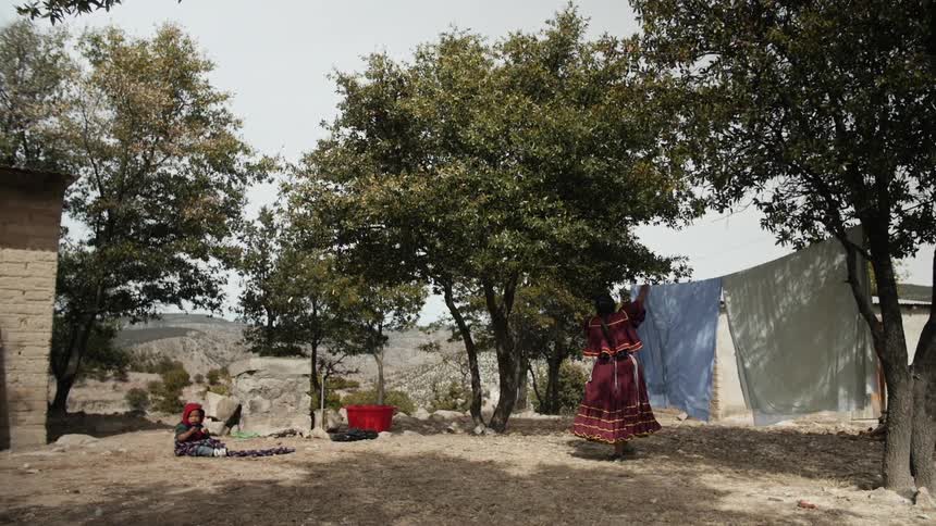 Woman hanging sheets outside on clothesline near child sitting in dirt