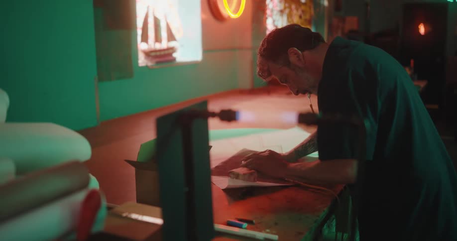 Man with a pencil in his mouth working on a project indoors with neon lights around him