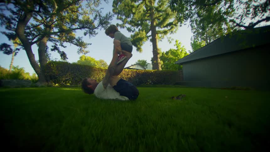 A man lying on his back in the grass lifting a young boy onto his legs